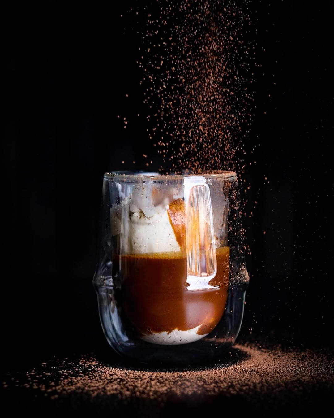 Affogato in a glass on a black ground with cocoa dust falling into the cup.