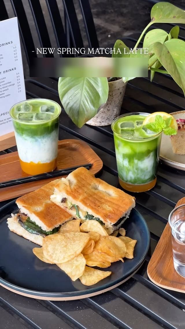 Spring matcha latte beside a toasted focaccia sandwich and crisps, with the menu board behind.